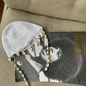 Può includere: Un cappello bianco all'uncinetto con finiture di conchiglie, appoggiato su una rivista in bianco e nero con la scritta dorata "LEGENDS". Il cappello è realizzato in un materiale a rete ed è decorato con diverse piccole conchiglie.