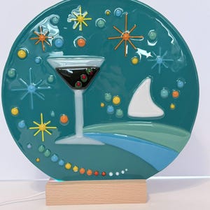 May include: A teal-colored, round decorative piece featuring a martini glass with olives, starbursts, and a sailboat design. The artwork is mounted on a wooden base with a light. The piece has a mid-century modern aesthetic.