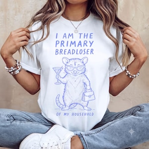 Op de afbeelding: Een wit T-shirt met korte mouwen en een blauwe afbeelding van een opossum met een zonnebril, die een martiniglas en een handtas vasthoudt. De tekst erboven luidt I AM THE PRIMARY BREADLOSER en de tekst eronder OF MY HOUSEHOLD in het blauw.