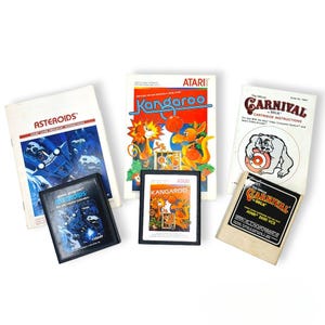 Puede incluir: Colección de cartuchos de videojuegos Atari antiguos y manuales de instrucciones. Los títulos incluyen "Asteroids", "Kangaroo" y "Carnival". Los cartuchos son negros y los manuales presentan gráficos y texto coloridos.