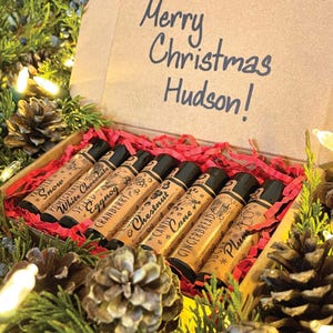 May include: A gift box with the text "Merry Christmas Hudson!" contains eight small, cylindrical bottles with black caps. Each bottle has a different flavor label, including "Vanilla Snow" and "Gingerbread". The box is surrounded by pine needles and pine cones.