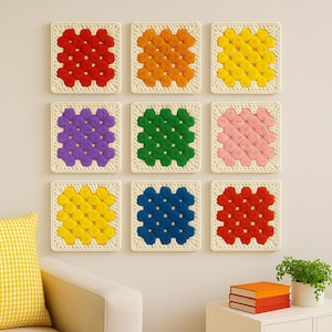 May include: Nine colorful crochet squares in a grid pattern. Each square features a different color: red, orange, yellow, purple, green, pink, yellow, blue, and red. The squares are mounted on a cream-colored background, creating a vibrant wall art display.