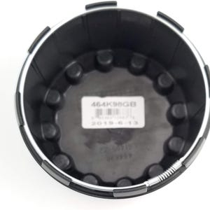 Pu&ograve; includere: Componente automobilistico in plastica nera con finiture argentate. La parte presenta un design circolare con elementi circolari in rilievo e un'etichetta con il testo "464K98GB" e "2019-6-13".