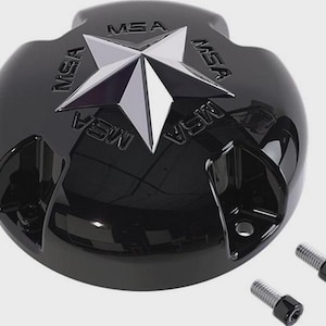 Puede incluir: Un componente negro brillante con un emblema de estrella plateada y las letras "MSA" en relieve alrededor del borde. Dos tornillos plateados con cabezas negras están colocados a la derecha. El objeto tiene una forma redondeada con una parte superior plana.