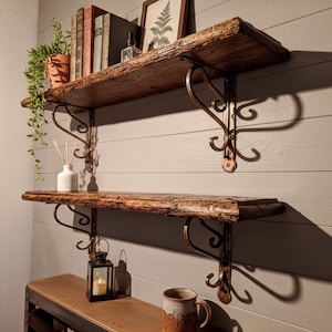 Könnte beinhalten: Zwei rustikale Holzregale mit verzierten, bronzefarbenen Metallhalterungen sind an einer grauen Wand befestigt. Das obere Regal enthält Bücher, einen gerahmten Druck und eine Topfpflanze. Das untere Regal hat eine Kerzenlaterne und einen Keramikbecher.