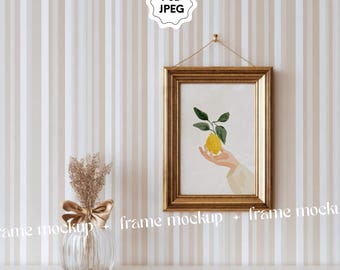 Op de afbeelding: Een goudomlijste kunstdruk met een hand die een citroen met groene bladeren vasthoudt. De print hangt aan een gestreepte muur boven een glazen vaas met gedroogde bloemen en een gouden strik. De tekst "PSD JPEG" is inbegrepen.