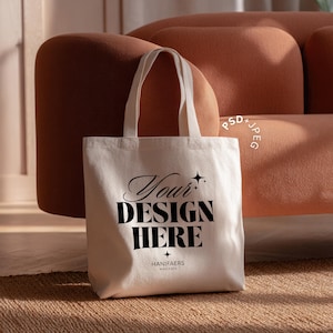Könnte beinhalten: Cremefarbene Tragetasche mit dem Text "Your DESIGN HERE" in schwarzer, eleganter Schrift. Die Tasche hat zwei Henkel und steht auf einem gewebten Teppich vor einem braunen Sofa. Die Worte "HANIFAERS MOCKUPS" stehen unten.