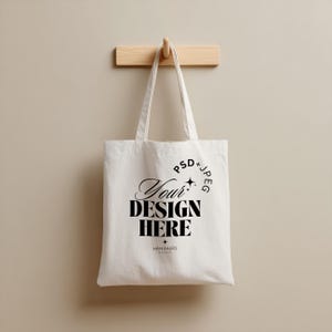 Peut inclure: Un sac fourre-tout en toile blanche avec les mots "Your Design Here" en écriture noire et en lettres majuscules. Le sac est suspendu à un crochet en bois sur un mur de couleur neutre. Le texte "PSD + JPEG" est également imprimé.