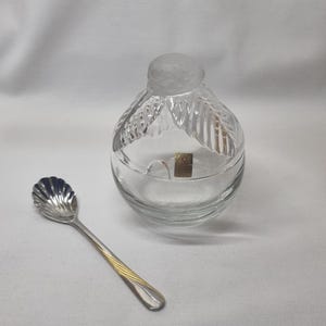 Könnte beinhalten: Ein klares Glasgefäß mit mattiertem Deckel und ein silberner Löffel mit goldenem Akzent. Das Glas hat ein dekoratives Blättermuster und ein kleines goldenes Etikett. Der Löffel hat eine muschelförmige Schale.