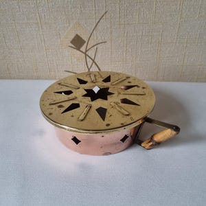 Könnte beinhalten: Ein Vintage-Räuchergefäß aus Kupfer und Messing. Der Deckel hat ein sternförmiges Design und ausgeschnittene Formen. Es hat einen Holzgriff. Der Brenner steht auf einer weißen Oberfläche.