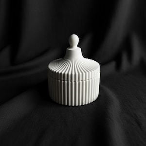 May include: A white ceramic jar with a lid and a small spherical handle. The jar has a cylindrical shape with vertical grooves and sits on a black fabric surface. The lid features a fluted design.