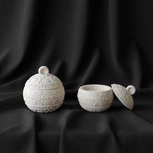 Peut inclure: Bougeoir en céramique blanche avec couvercle et bougeoir ouvert séparé. Les deux sont décorés de motifs de flocons de neige et géométriques. Le couvercle a une petite poignée en boucle. Le bougeoir est sur fond noir.