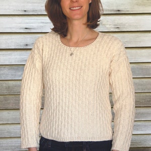 May include: A woman wearing a cream-colored knitted sweater with a cable knit pattern and blue jeans. She is standing in front of a wood panel background.