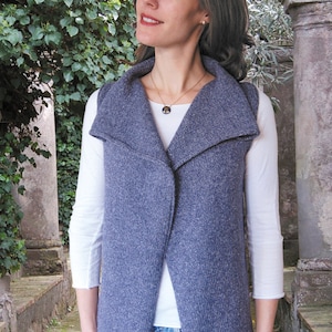 May include: A woman wearing a gray knit vest with a wide collar and a white long-sleeved shirt underneath. She is wearing blue jeans and a silver necklace with a round pendant.