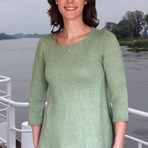 May include: A woman wearing a light green knitted sweater with a scoop neck and three-quarter sleeves. She is standing in front of a body of water and a white railing.