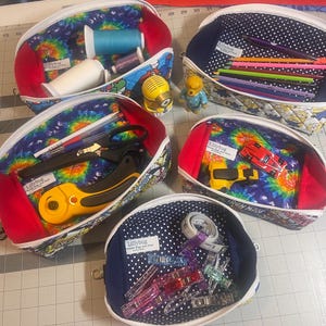 May include: Several open zippered pouches with various contents. The pouches have a red interior and a white zip. The interiors feature tie-dye, polka dot, and comic book fabric. Contents include sewing supplies, pencils, and small toys. The label "Lillybug" is visible.