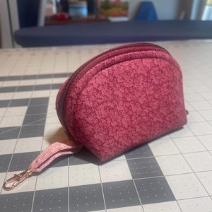 Może przedstawiać: Mała, półokrągła kosmetyczka na zamek błyskawiczny w głębokim różowym wzorze kwiatowym. Kosmetyczka ma pasujący różowy pasek na nadgarstek z zapięciem w kolorze złotym. Tło to szaro-biała mata do cięcia.