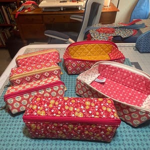 May include: A collection of quilted fabric pouches in various sizes and shapes, featuring floral and geometric patterns in shades of red, pink, yellow, and white. The pouches have zippered closures and some are open to reveal interior linings.