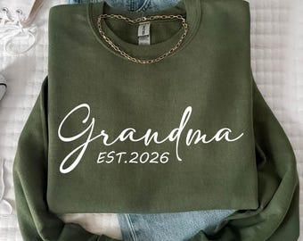 May include: An olive green sweatshirt with the word "Grandma" in white script and the text "EST. 2026". The sweatshirt is paired with light blue jeans, white high-top sneakers, and a gold chain necklace.