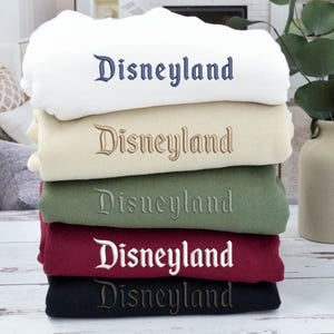 May include: A stack of folded sweatshirts in various colors: white, beige, green, burgundy, and black. Each sweatshirt has the word "Disneyland" embroidered across the front in a complementary color.
