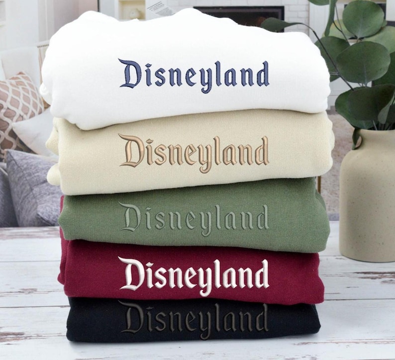 May include: A stack of folded sweatshirts in various colors: white, beige, green, burgundy, and black. Each sweatshirt has the word "Disneyland" embroidered across the front in a complementary color.