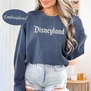 May include: A blue crew neck sweatshirt with the word "Disneyland" embroidered in white script. The sweatshirt has long sleeves and is paired with denim shorts. The word "Embroidered" is in a blue oval in the upper left corner.
