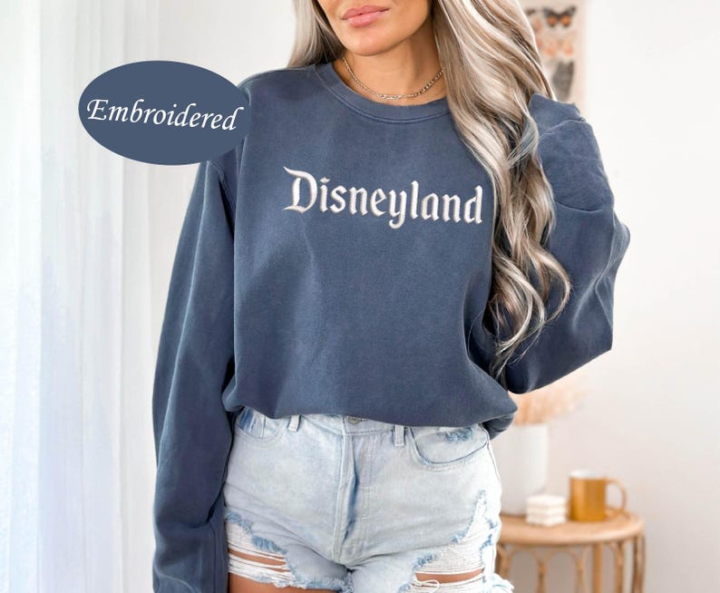 May include: A blue crew neck sweatshirt with the word "Disneyland" embroidered in white script. The sweatshirt has long sleeves and is paired with denim shorts. The word "Embroidered" is in a blue oval in the upper left corner.
