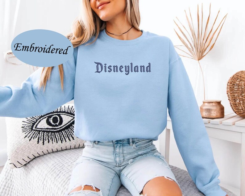 May include: A light blue sweatshirt with the word "Disneyland" embroidered in a dark blue script. The word "Embroidered" is in a light blue oval in the upper left corner. The sweatshirt is long-sleeved.
