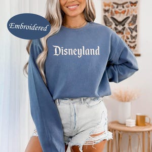 May include: A blue long-sleeve sweatshirt with the word "Disneyland" embroidered in white script. The sweatshirt is a solid blue color and has a relaxed fit. The word "Embroidered" is in a blue oval in the upper left corner.