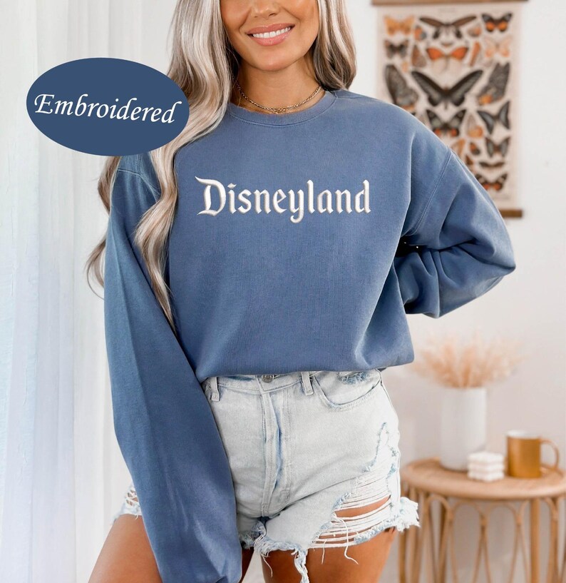 May include: A blue long-sleeve sweatshirt with the word "Disneyland" embroidered in white script. The sweatshirt is a solid blue color and has a relaxed fit. The word "Embroidered" is in a blue oval in the upper left corner.