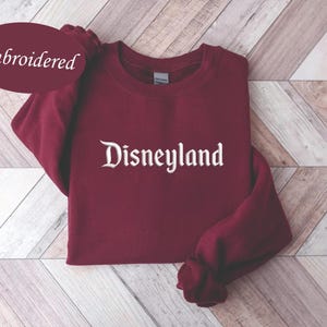 May include: A maroon sweatshirt with the word "Disneyland" embroidered in white script. The word "Embroidered" is in a maroon oval in the upper left corner. The sweatshirt is folded on a light brown and white wooden surface.