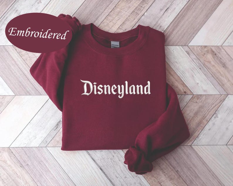 May include: A maroon sweatshirt with the word "Disneyland" embroidered in white script. The word "Embroidered" is in a maroon oval in the upper left corner. The sweatshirt is folded on a light brown and white wooden surface.