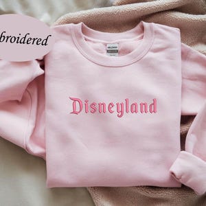 May include: A light pink sweatshirt with the word "Disneyland" embroidered in a matching pink script. The word "Embroidered" is in a light pink oval in the upper left corner. The sweatshirt has long sleeves and a crew neck.