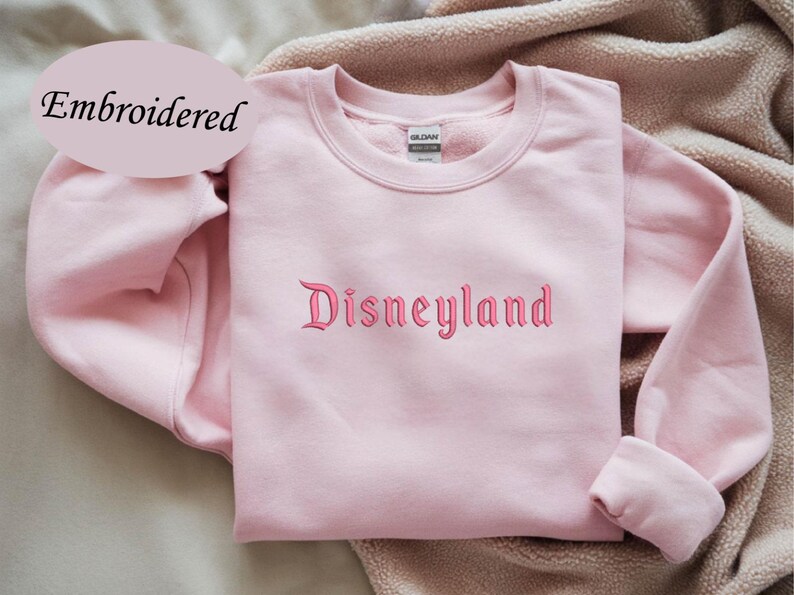 May include: A light pink sweatshirt with the word "Disneyland" embroidered in a matching pink script. The word "Embroidered" is in a light pink oval in the upper left corner. The sweatshirt has long sleeves and a crew neck.