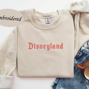 May include: A beige sweatshirt with the word "Disneyland" embroidered in red script across the chest. The sweatshirt has long sleeves and a crew neck. The word "Embroidered" is above the sweatshirt. The sweatshirt is paired with blue denim shorts and a brown mug.