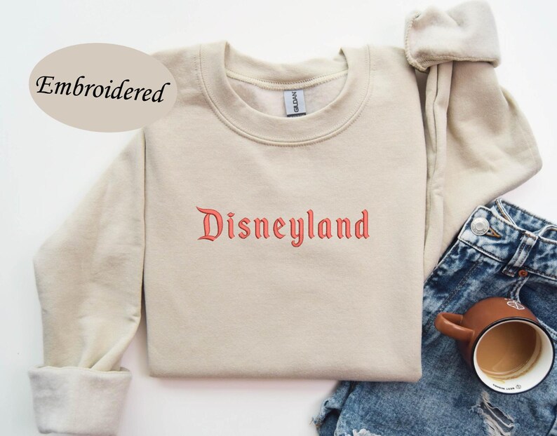 May include: A beige sweatshirt with the word "Disneyland" embroidered in red script across the chest. The sweatshirt has long sleeves and a crew neck. The word "Embroidered" is above the sweatshirt. The sweatshirt is paired with blue denim shorts and a brown mug.
