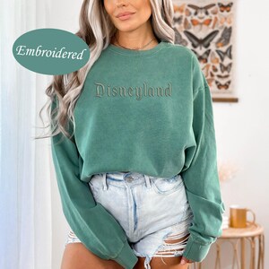 May include: A teal green sweatshirt with the word "Disneyland" embroidered across the chest. The sweatshirt has long sleeves and a crew neck. The word "Embroidered" is in a teal green oval in the upper left corner.
