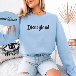 May include: A light blue sweatshirt with the word "Disneyland" embroidered in black gothic font. The word "Embroidered" is in a blue oval on the left side of the image. The sweatshirt has long sleeves.