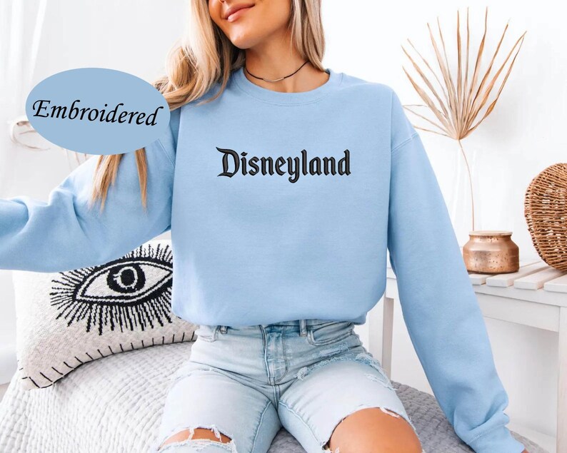May include: A light blue sweatshirt with the word "Disneyland" embroidered in black gothic font. The word "Embroidered" is in a blue oval on the left side of the image. The sweatshirt has long sleeves.