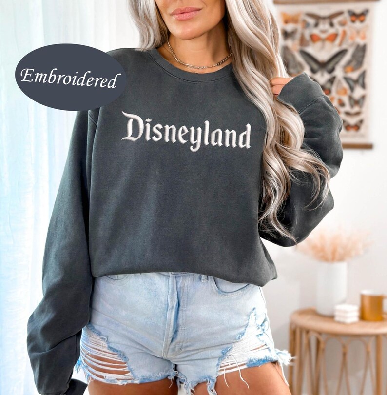 May include: Dark gray embroidered sweatshirt with the word "Disneyland" in white script. The sweatshirt has long sleeves and a crew neck. The word "Embroidered" is above the word "Disneyland".