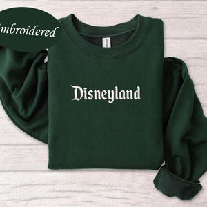 May include: A dark green sweatshirt with the word "Disneyland" embroidered in white script across the chest. The word "Embroidered" is in a dark green oval in the upper left corner. The sweatshirt is folded on a light wood surface.