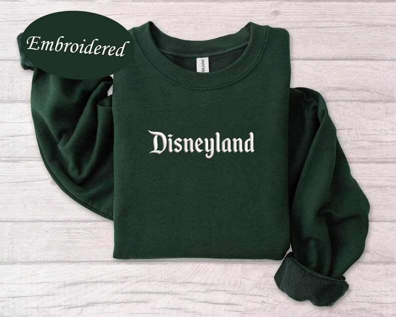 May include: A dark green sweatshirt with the word "Disneyland" embroidered in white script across the chest. The word "Embroidered" is in a dark green oval in the upper left corner. The sweatshirt is folded on a light wood surface.