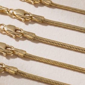 May include: Close-up of several gold-colored snake chain necklaces. Each necklace features a delicate, textured chain and a secure clasp. The chains are arranged parallel, showcasing their design and the warm, metallic sheen of the gold.