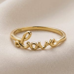 May include: A gold ring with the word "love" in a cursive script. The ring is made of 14K gold and sits on a cream-colored fabric. The ring is a delicate piece of jewelry.