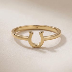 May include: A gold-colored ring with a horseshoe design. The ring is made of 14K gold and is set against a neutral background. The horseshoe is a classic symbol of luck and good fortune.