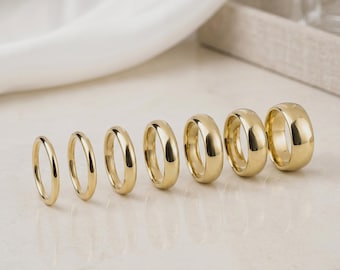 May include: A collection of seven gold wedding bands of varying widths, displayed on a light-colored surface. The rings are arranged from thinnest to thickest, showcasing their polished, reflective surfaces. The background includes a white fabric and a perfume bottle.