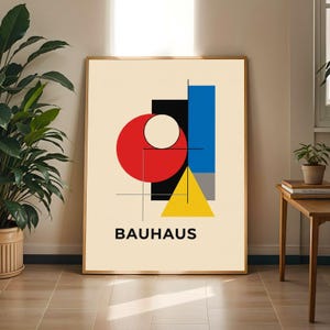 May include: A framed Bauhaus art print with a cream background, displaying geometric shapes in red, black, blue, yellow, and grey. The word "BAUHAUS" is printed in black at the bottom. The artwork is displayed in a room with a wooden floor and a potted plant.