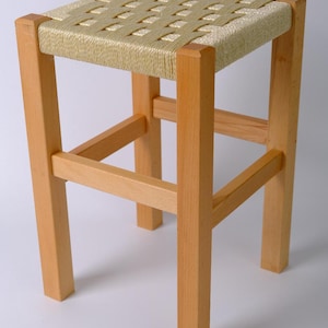 Puede incluir: Un taburete de madera clara con un asiento tejido. El asiento cuadrado es de un material beige claro, y las patas son de color madera natural. El taburete tiene un diseño simple y minimalista, con barras transversales para mayor estabilidad. El taburete mide aproximadamente 46 cm de alto.