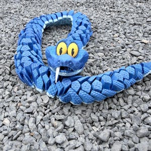 May include: A blue, segmented snake toy with yellow eyes and a white fang. The toy is posed on a bed of gray gravel. The snake's body is made of many small, textured pieces.