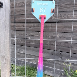 May include: A baseball bat and a light blue home plate-shaped sign with the name "Beckett" and the number "67" are displayed. The bat is pink, purple, and blue with the word "WARSTIC" printed on it. The sign is mounted on a wire fence.